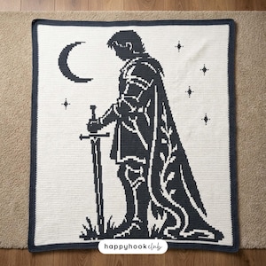 Puede incluir: Una manta de ganchillo rectangular con fondo blanco y borde azul marino oscuro. El diseño muestra la silueta de una figura con armadura sosteniendo una espada, acompañada por una luna creciente y varias estrellas de cuatro puntas.