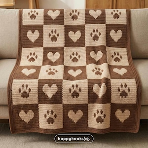 Może przedstawiać: Brązowo-beżowy koc szydełkowy z wzorem szachownicy z odciskami łap i sercami. Koc jest narzucony na sofę. Tekst "happyhookclub" znajduje się w prawym dolnym rogu.