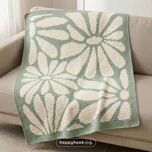 May include: A crocheted blanket with a daisy pattern in cream and light green. The blanket is folded over a beige sofa. The daisy design is a repeating pattern across the blanket. The blanket has a light green border.