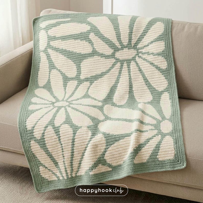 May include: A crocheted blanket with a daisy pattern in cream and light green. The blanket is folded over a beige sofa. The daisy design is a repeating pattern across the blanket. The blanket has a light green border.