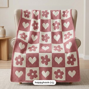 May include: A pink and white crocheted blanket draped over a chair. The blanket features a checkerboard pattern with alternating squares of flowers and hearts. The text "happyhookclub" is visible in the bottom right corner.