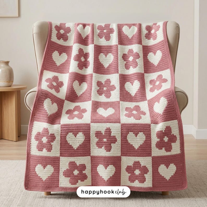May include: A pink and white crocheted blanket draped over a chair. The blanket features a checkerboard pattern with alternating squares of flowers and hearts. The text "happyhookclub" is visible in the bottom right corner.