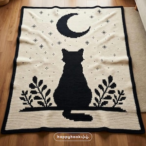 Pode incluir: Um cobertor de crochê creme com uma silhueta de gato preto sob uma lua crescente e estrelas. O cobertor tem uma borda preta e desenhos de folhas. O texto "happyhookclub" está no canto inferior direito.