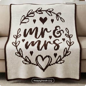 Puede incluir: Una manta de ganchillo color crema con un borde marrón. La manta presenta las palabras "mr & mrs" en una fuente decorativa, rodeadas de una corona de hojas y corazones. La manta está doblada sobre un sofá beige.