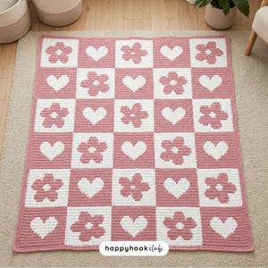 May include: A handmade, pink and white crochet blanket with a repeating pattern of flowers and hearts. The blanket is square and features a checkerboard design. The text "happyhook club" is visible in the bottom right corner.