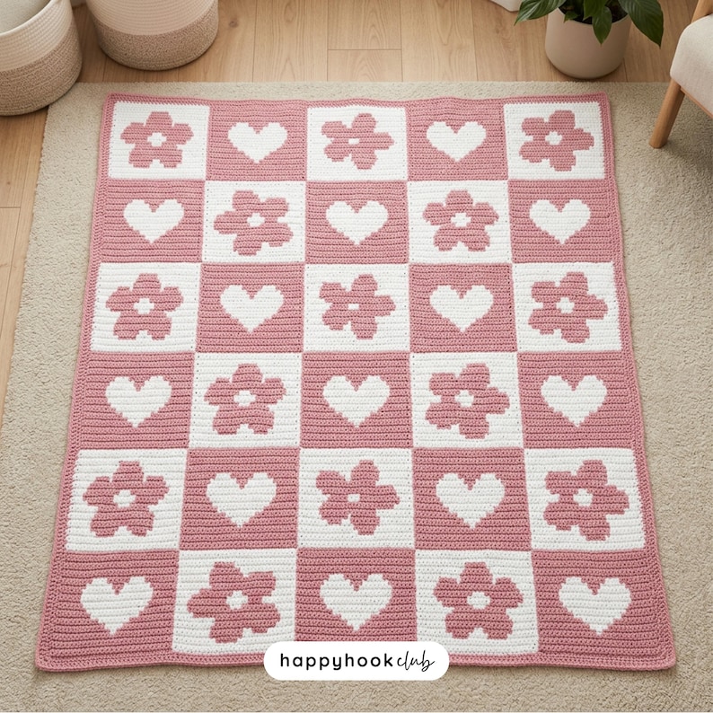 May include: A handmade, pink and white crochet blanket with a repeating pattern of flowers and hearts. The blanket is square and features a checkerboard design. The text "happyhook club" is visible in the bottom right corner.