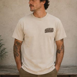 May include: A person wearing a brown baseball cap and a beige t-shirt with the text "THE RED CLAY STRAYS" printed on the left chest. The person is also wearing olive green pants.