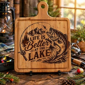 May include: A rectangular wooden cutting board with a handle and a black metal stand. The board features an engraved design of a fish jumping out of the water, mountains, trees, and the text "LIFE IS Better AT THE LAKE."