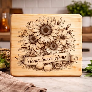 May include: A square wooden chopping board with a laser-etched design. The design features sunflowers, other flowers, and the text "Home Sweet Home" on a banner. Two eggs are also etched below the banner. The board has a light wood grain.