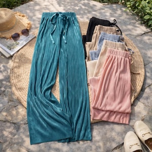 May include: Pleated wide-leg pants in teal, with a drawstring waist, are displayed alongside other colored pants. Additional colors include black, beige, light blue, and pink. The pants are arranged on a woven mat, with a straw hat and sunglasses visible.