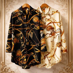 May include: Two long-sleeved button-down shirts hang on wooden hangers. One is black with a gold chain and floral pattern, the other cream-coloured with a similar gold chain and floral design. Both shirts have a classic, elegant style, suitable for various occasions.