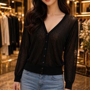 May include: A black, sheer cardigan with a V-neck and button-up front. The long-sleeved top has a fitted waist and cuffs. The cardigan is worn with blue jeans. The background shows clothing racks and decorative items.