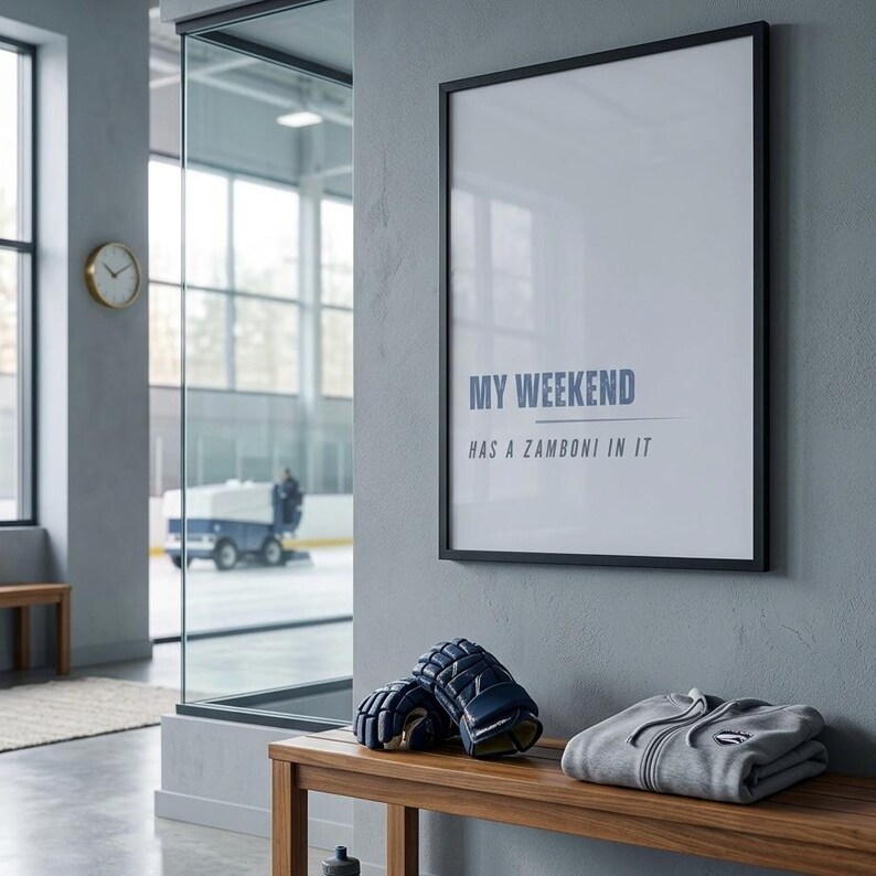 May include: A framed print with the text "MY WEEKEND HAS A ZAMBONI IN IT" hangs on a gray wall. Below, a wooden bench holds hockey gloves and a gray zip-up hoodie. A clock and a view of an ice rink are visible.