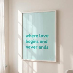 Könnte beinhalten: Ein gerahmter Druck mit hellblauem Hintergrund und weißem Rahmen. Der Text "where love begins and never ends" wird in türkisfarbenen Buchstaben angezeigt. Der Druck hängt an einer weißen Wand.