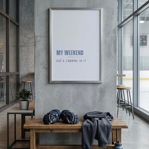 May include: A framed poster with the text "MY WEEKEND HAS A ZAMBONI IN IT" hangs on a concrete wall. Below, a wooden bench holds hockey gloves and a gray sweatshirt. A small potted plant and stools are also visible.