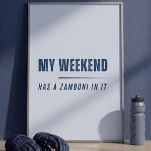 May include: Framed print with the text "MY WEEKEND HAS A ZAMBONI IN IT" in navy blue. A gray water bottle and hockey gloves are visible. The background is a solid navy blue.