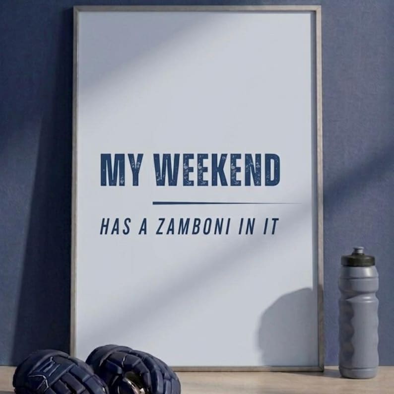 May include: Framed print with the text "MY WEEKEND HAS A ZAMBONI IN IT" in navy blue. A gray water bottle and hockey gloves are visible. The background is a solid navy blue.