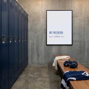 May include: A locker room scene with navy blue lockers, a wooden bench, and a framed print. The print reads "MY WEEKEND HAS A ZAMBONI IN IT." A jersey, gloves, and a towel rest on the bench. The room has a concrete floor and wall.