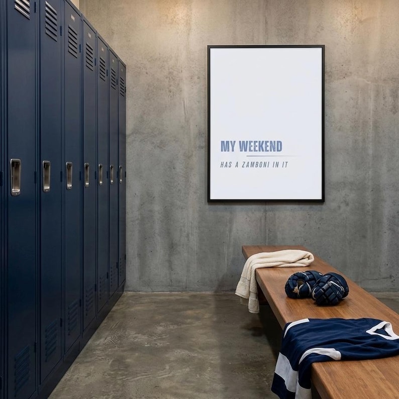 May include: A locker room scene with navy blue lockers, a wooden bench, and a framed print. The print reads "MY WEEKEND HAS A ZAMBONI IN IT." A jersey, gloves, and a towel rest on the bench. The room has a concrete floor and wall.