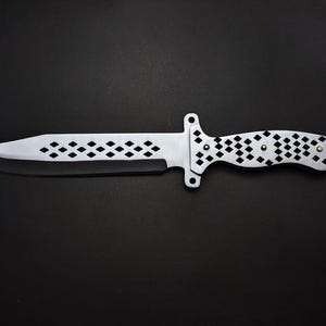 Może przedstawiać: Srebrny nóż z czarnym ostrzem, z rękojeścią i ostrzem ozdobionymi wycięciami w kształcie rombów. Nóż ma osłonę i jest umieszczony na czarnym tle.