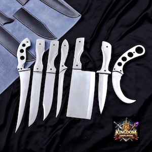 Può includere: Un set di sette coltelli in acciaio inossidabile con manici bianchi, tra cui un'accetta e un coltello karambit curvo. I coltelli sono esposti su una superficie nera con una custodia blu e grigia sullo sfondo. Il logo "KINGDOM COSPLAYERS" è visibile.
