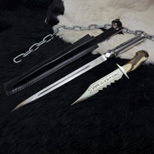 May include: A silver dagger with a serrated edge and a deer antler handle, resting on a black surface. The dagger is accompanied by a black leather sheath and a silver chain.