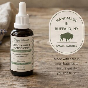 ​A product shot of an amber glass dropper bottle of Peppy Momma GHK-Cu & SNAP-8 Peptide Serum resting on a rustic wooden surface next to dried lavender. To the right of the bottle is an oval badge with a buffalo silhouette that reads "Handmade in Buffalo, NY, Small Batches." Below the badge, text reads, "Made with care in small batches to ensure quality you can trust."