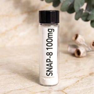 May include: A clear glass vial with a black screw-top lid containing a white powder. A white rectangular label is affixed to the front of the vial with the black printed text SNAP-8 100mg. The vial stands upright in a vertical orientation.