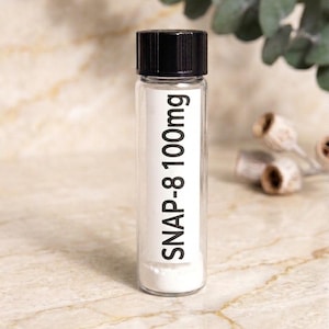 May include: A clear glass vial with a black screw-top lid containing a white powder. A white rectangular label is affixed to the front of the vial with the black printed text SNAP-8 100mg. The vial is cylindrical and stands upright.