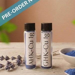 Puede incluir: Dos pequeños frascos de vidrio con tapas negras, etiquetados como "AHK-Cu 1g" y "GHK-Cu 1g", contienen polvo azul. Un pequeño cuenco blanco con polvo azul y ramitas de lavanda también están presentes. El texto dice "PRE-ORDER NOW" y "SECURE YOURS! PRE-ORDER OPEN."