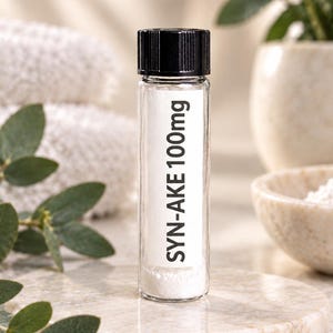 May include: A clear glass vial with a black screw-top lid containing a white powder. A white rectangular label on the front of the vial features the black printed text SYN-AKE 100mg. The vial is cylindrical and transparent, showing the contents.