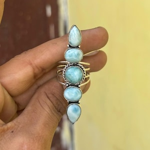 Könnte beinhalten: Ein Ring aus Sterlingsilber mit fünf vertikal angeordneten, ovalen, hellblauen Larimar-Edelsteinen. Die Steine sind in glatten Fassungen eingefasst, wobei der mittlere Stein von einem gedrehten Metalldetail und einem mehrbändigen Schaft umgeben ist.