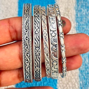 Könnte beinhalten: Ein Set aus fünf silberfarbenen Metallarmreifen mit verschiedenen geprägten Mustern. Die Designs umfassen verschlungene Ranken, ein florales Motiv, ein zartes Blattmuster, ein Himmelsmotiv mit Sternen und Monden sowie einen Perlenrand.
