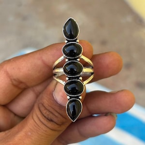 Puede incluir: Un anillo de plata con cinco piedras negras lisas, de forma ovalada y de gota, alineadas verticalmente. La banda de metal se divide en varios hilos de plata pulida que se conectan a los engastes de las piedras, creando un diseño alargado.