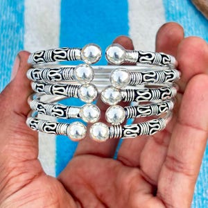 Könnte beinhalten: Ein silberfarbenes Armband bestehend aus fünf gestapelten Reifen. Jeder Reifen ist an beiden Enden mit einer polierten, kugelförmigen Perle versehen und weist ein sich wiederholendes, schwarz oxidiertes Muster auf dem Metall auf.