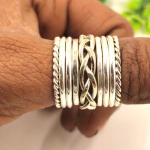 Könnte beinhalten: Eine Ansammlung von Silberringen an einem Finger. Die Ringe umfassen glatte Bänder, geflochtene Designs und verdrehte, seilartige Texturen. Die Ringe sind übereinander gestapelt, wodurch ein geschichteter Effekt entsteht. Die Ringe bestehen aus glänzendem Metall.