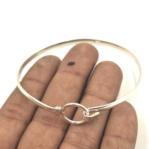 Könnte beinhalten: Ein silbernes Armreif-Armband mit Haken- und Ösenverschluss. Das Armband ist dünn und rund, mit einem schlichten, eleganten Design. Der Verschluss ist eine kreisförmige Schlaufe, die mit einem gebogenen Haken verbunden ist. Das Armband wird auf einer Hand gezeigt.
