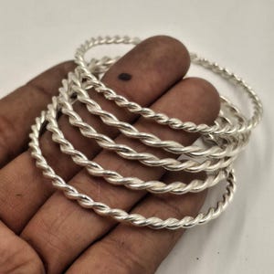 Könnte beinhalten: Eine Sammlung silberfarbener Armreifen im gedrehten Seil-Stil. Die Armreifen sind übereinander gestapelt, wobei die Details des gedrehten Designs deutlich sichtbar sind. Der Schmuck wird vor einem neutralen Hintergrund präsentiert.