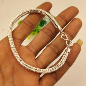 May include: A silver-colored bracelet with a woven chain design. The bracelet is held in a hand, showcasing its intricate pattern and the secure clasp. The bracelet is a piece of jewelry.