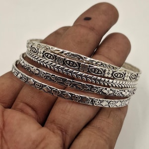 Könnte beinhalten: Ein Set aus vier silbernen Armreifen mit aufwendigen Blumen- und Blättermustern. Die Armreifen sind gestapelt und zeigen die detaillierten Designs. Der Schmuck wird auf einer Hand präsentiert, um die Handwerkskunst und den Stil hervorzuheben.