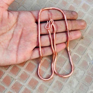 Könnte beinhalten: Eine kupferfarbene Schlangenkette mit einem glatten, flexiblen und segmentierten Metalldesign. Die Kette ist mit einem einfachen Hakenverschluss versehen und weist über die gesamte Länge eine polierte, reflektierende Oberfläche auf.