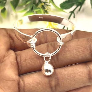 Könnte beinhalten: Ein silberfarbenes Armband mit einem kreisförmigen Mittelstück und einem tropfenförmigen Anhänger. Das Armband ist schlicht und elegant, mit poliertem Finish. Der Anhänger hängt frei.