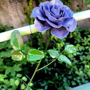 May include: A decorative flower sculpture featuring a single, multi-layered purple rose with a glittery, shimmering finish. The rose is attached to a thin, green metal stem that includes several glossy, vibrant green leaves extending outward.