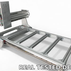 May include: A silver CNC machine with a rectangular frame and a gantry system. The machine has a series of parallel rails and a vertical support structure. The text "REAL TESTED DESIGN" is displayed at the bottom.