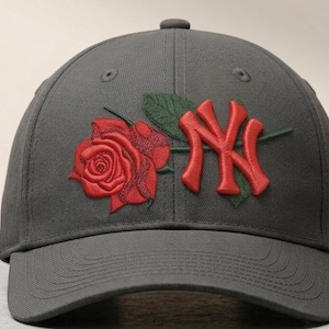Puede incluir: Gorra de béisbol gris con una rosa roja bordada y el logotipo de los Yankees de Nueva York en rojo. La gorra tiene una visera curva y un botón en la parte superior. La rosa está detallada con hojas verdes.