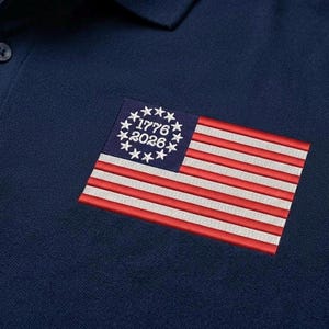 May include: Navy blue polo shirt with a small embroidered American flag patch. The flag features red and white stripes, a blue field with stars, and the dates "1776" and "2026". The shirt has a classic collar and button placket.