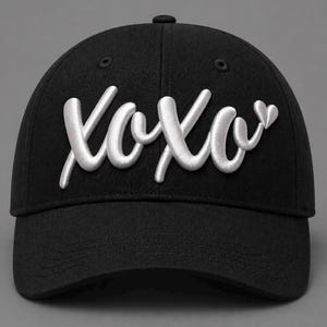 May include: A black baseball cap featuring the white embroidered text XOXO in a cursive script font across the front panels. The design includes a small white embroidered heart shape positioned to the right of the final letter O.