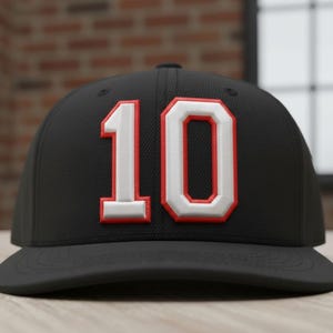 Könnte beinhalten: Schwarze Baseballkappe mit der Zahl "10" in Weiß mit roter Umrandung. Die Kappe hat einen flachen Schirm und eine strukturierte Krone. Die Zahl ist ein wichtiges Designelement auf der Vorderseite.
