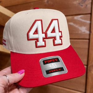Könnte beinhalten: Beige und rote Baseballkappe mit der weißen und roten Zahl "44". Die Kappe hat einen roten Schirm und ein kleines Abzeichen mit den Wörtern "POPES" und "FERICH".