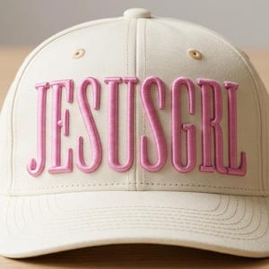 May include: A cream-colored baseball cap with the embroidered text "JESUSGRL" in pink. The cap features a curved brim, a button on top, and stitched details.
